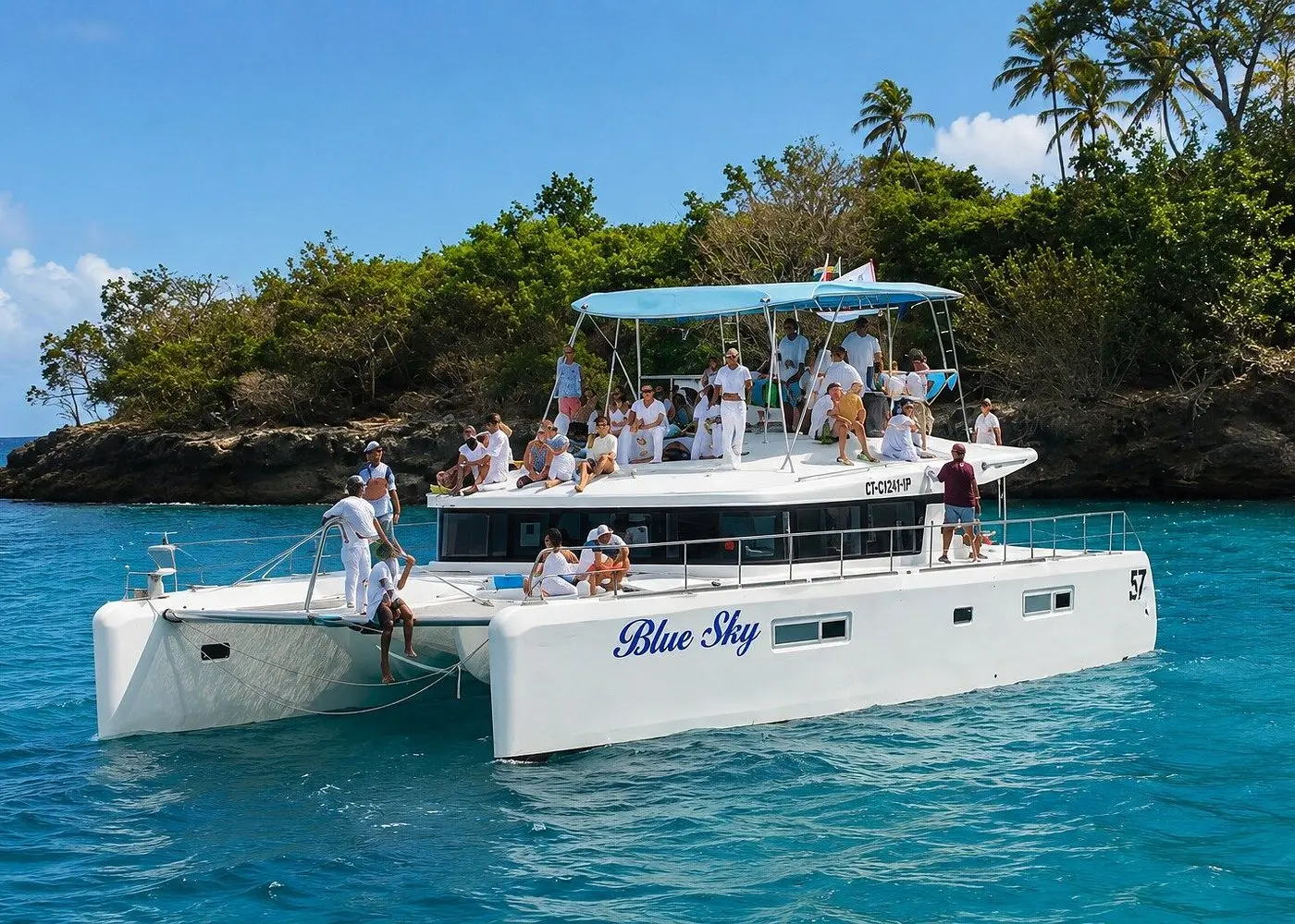 Guests on the decks of a white catamaran anchored by a rocky, palm-topped headland off Puerto Plata