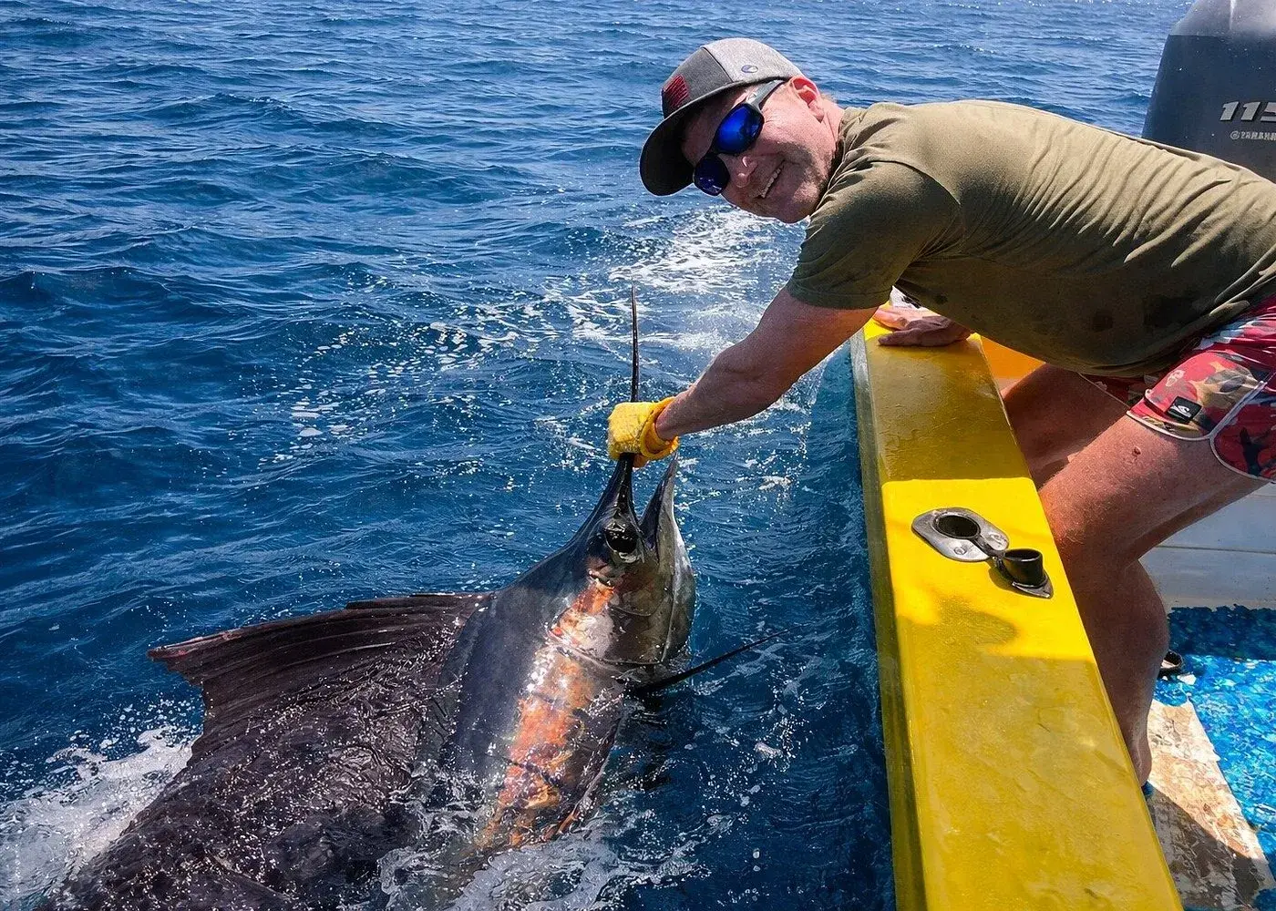 A smiling angler aboard a fishing boat carefully releases a large sailfish into the ocean during an offshore sport fishing trip in deep blue tropical waters.