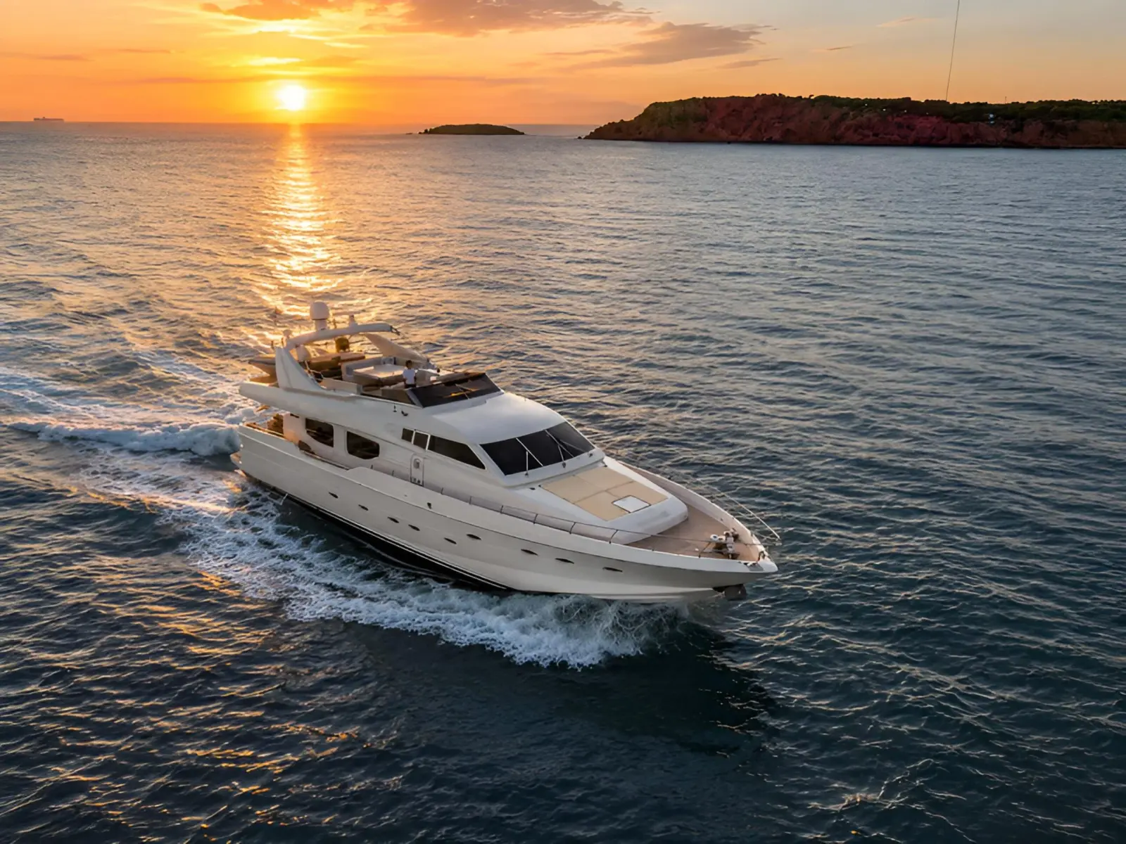 Luxury private yacht cruising across the ocean at sunset, with golden reflections on the water and a rocky coastline in the background.