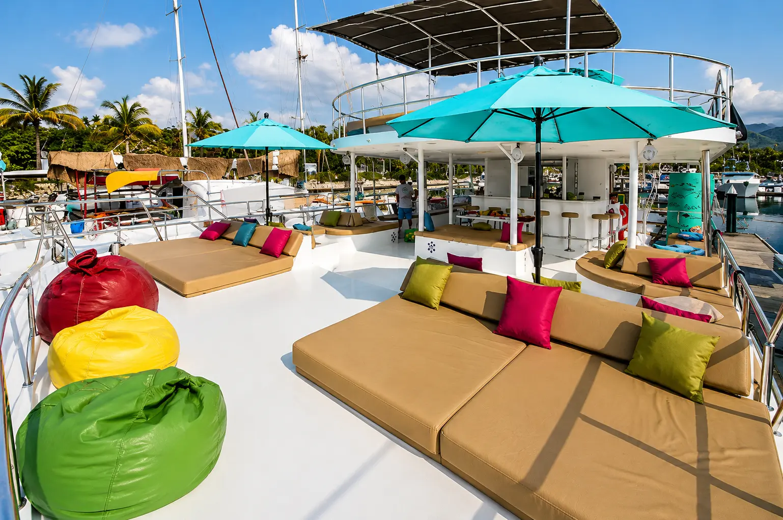 The catamaran's sun deck with cushioned lounges, bright beanbags and teal umbrellas, docked at the marina