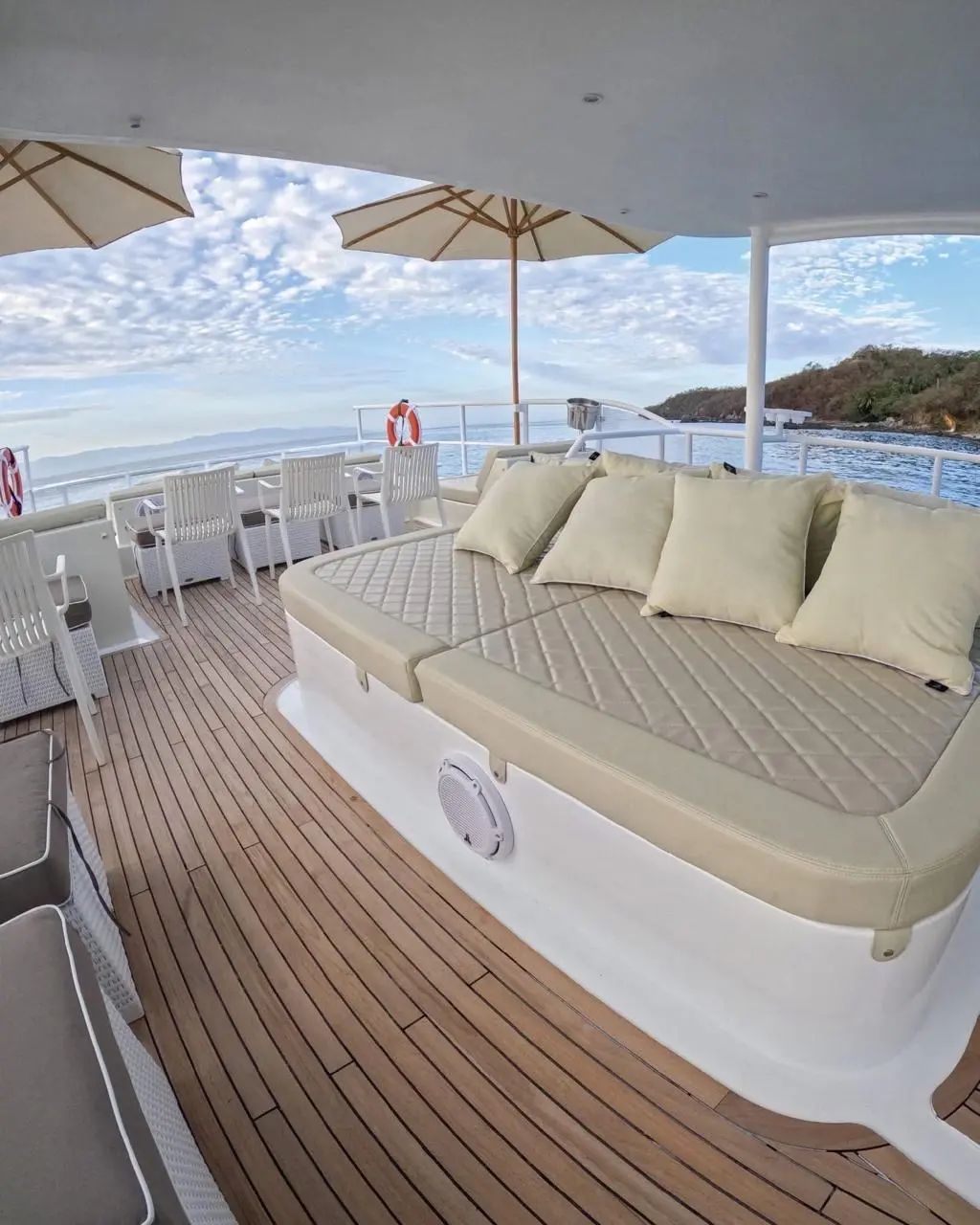 Padded sunbed with pillows on the catamaran's teak deck, parasols and a green headland beyond
