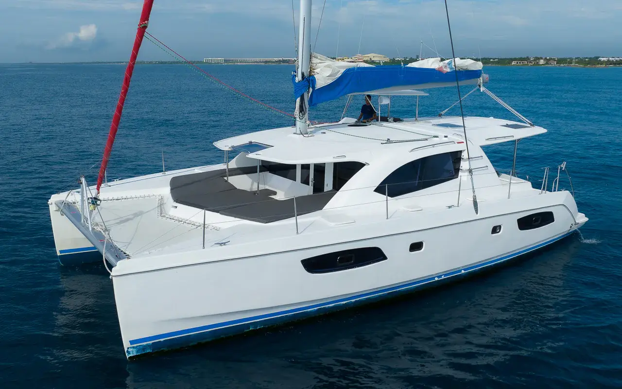 Sailing catamaran at anchor with sails furled on calm blue water, the low Riviera Maya coastline beyond