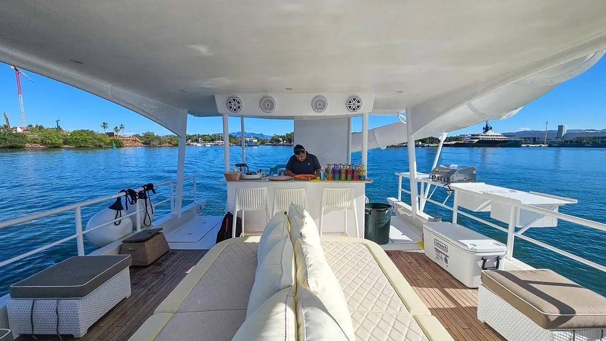 A crew member tends the bar on the catamaran's shaded upper deck, cushioned seats and the bay beyond