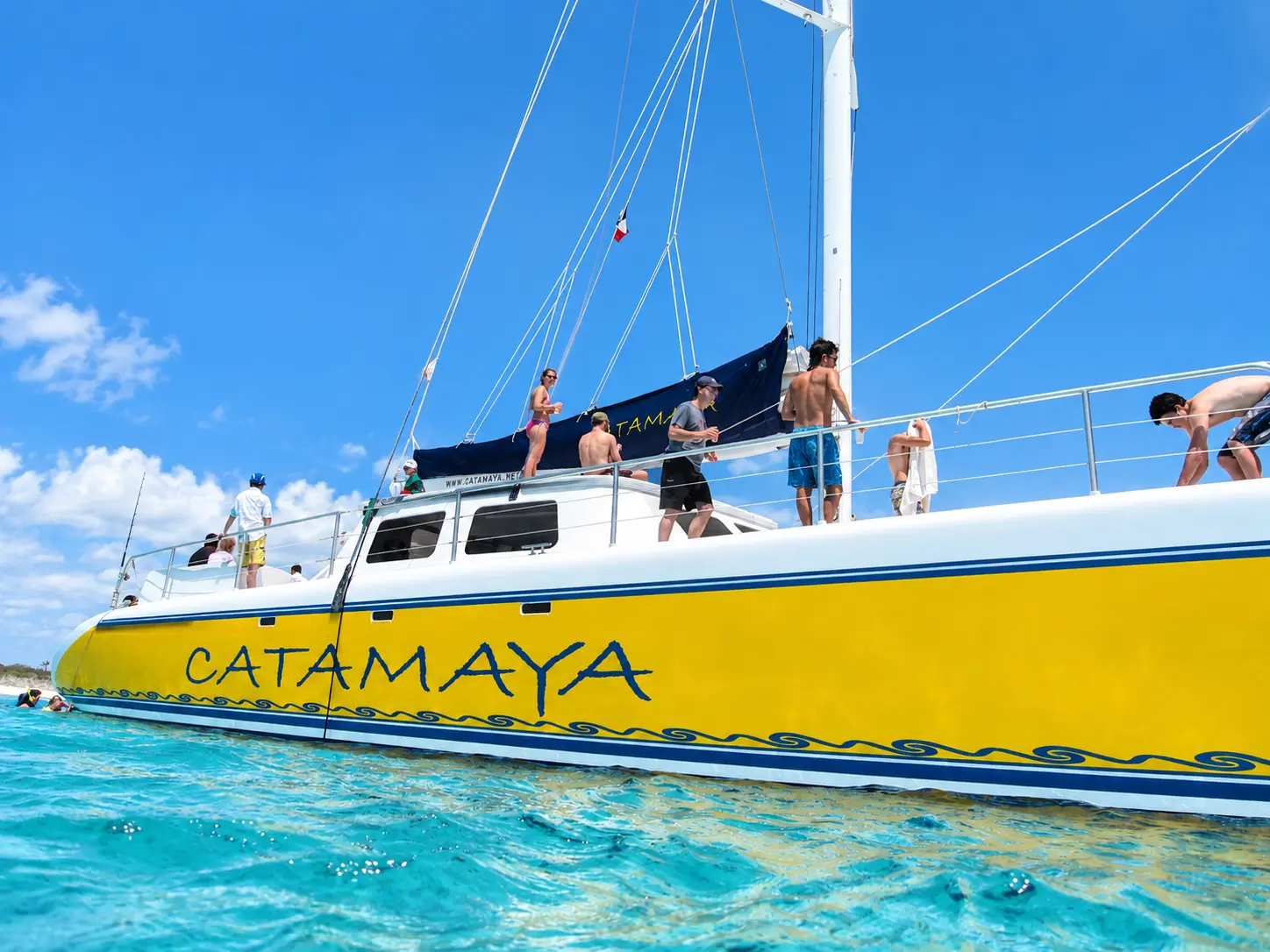 Low view of the yellow sailing catamaran's hull with guests on deck above turquoise Riviera Maya shallows