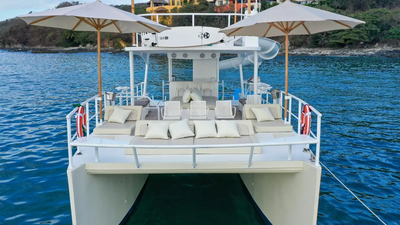 Two-level catamaran's bow with cushioned loungers and parasols, anchored off a green Puerto Vallarta shore