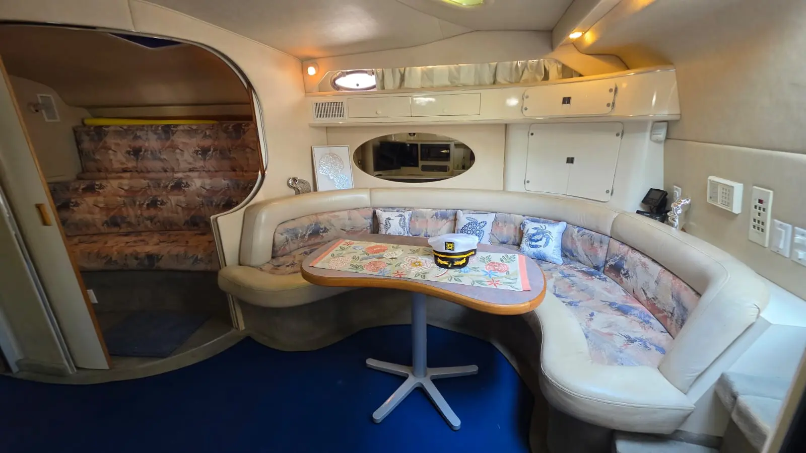Yacht cabin with a curved floral-print dinette, a captain's hat on the table and blue carpet