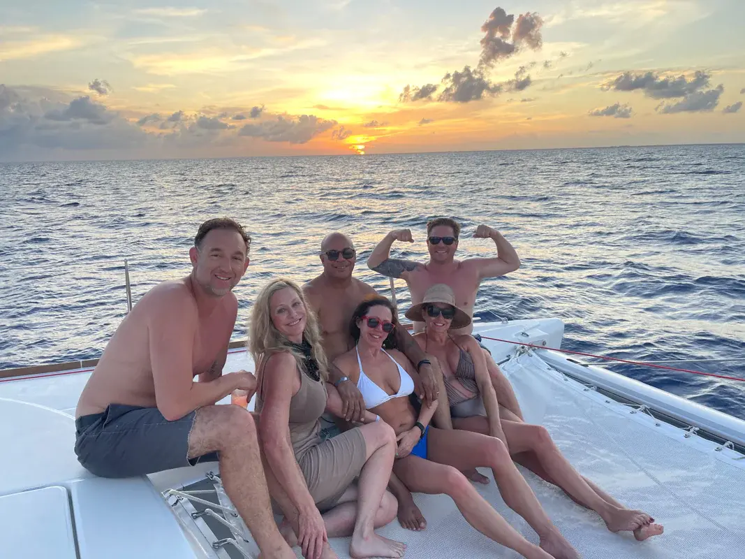 Friends posing on the catamaran's bow at sunset, one man flexing his arms