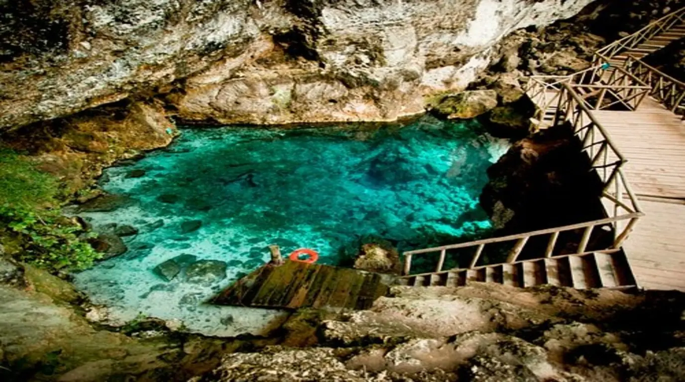 Wooden stairs leading down to a turquoise cenote pool beneath a limestone overhang near Punta Cana
