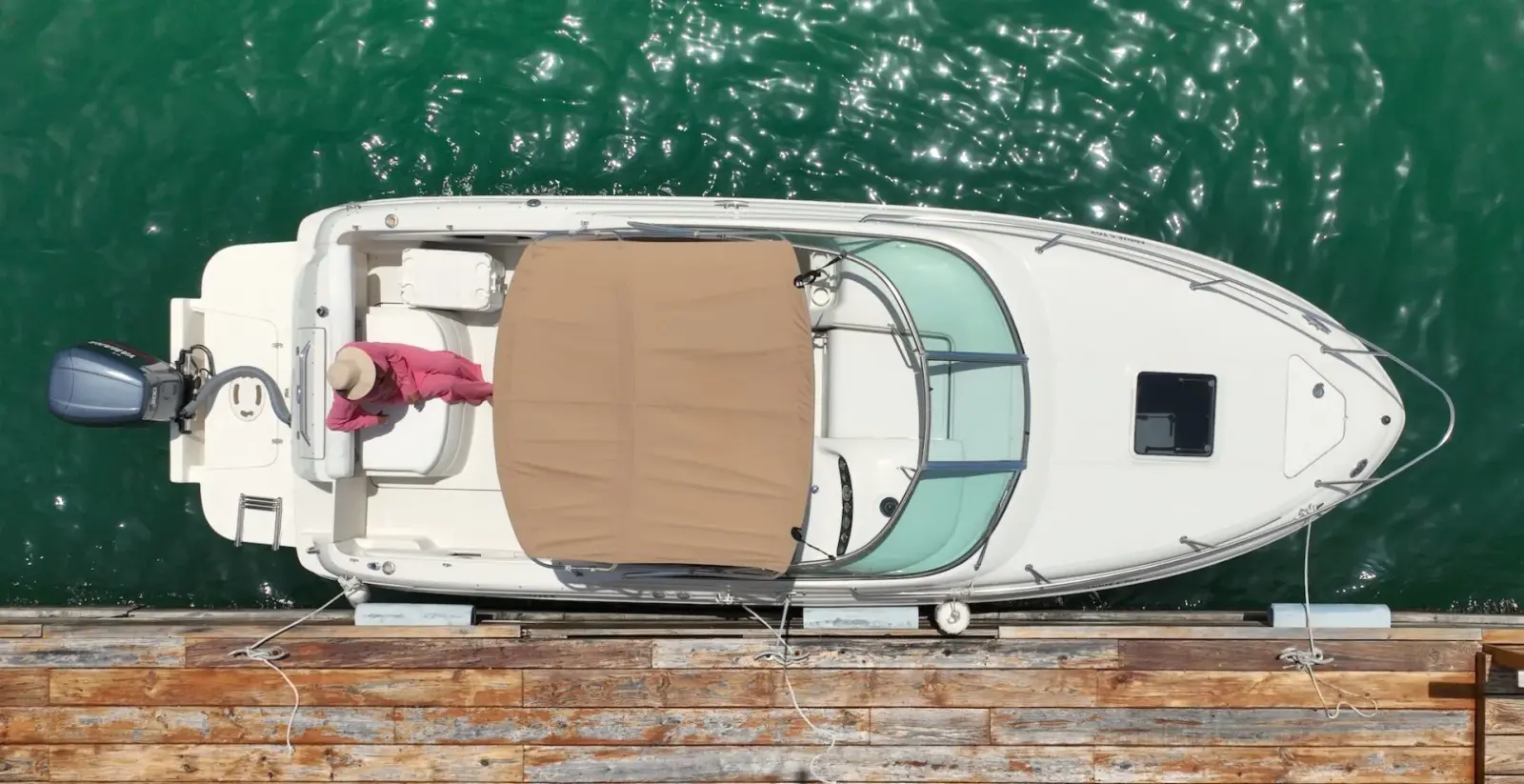 Top-down aerial view of a private white motorboat docked at a marina with emerald-green water, wooden pier, and a person on board preparing for a relaxing boat excursion.