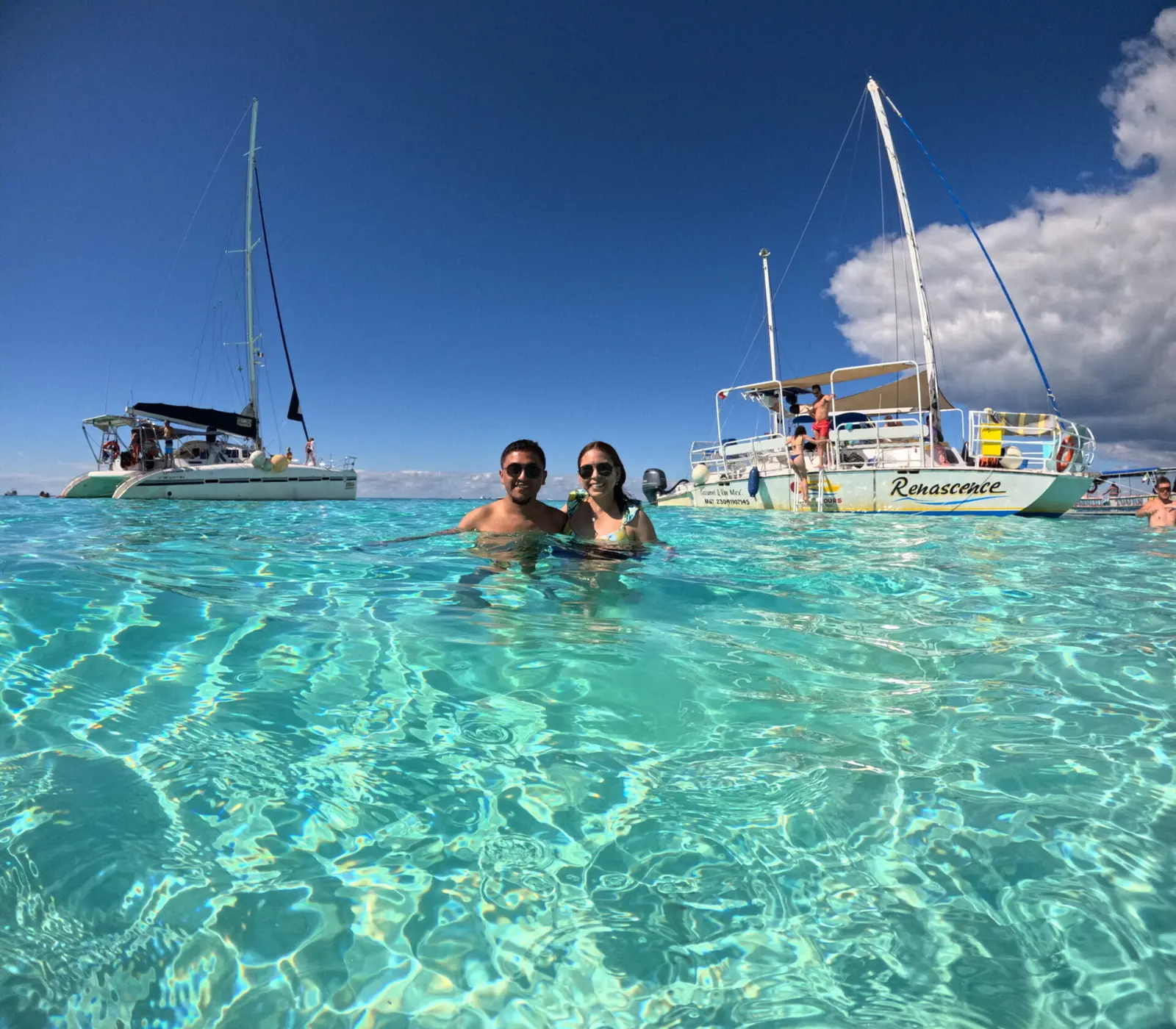 Couple standing chest-deep in turquoise shallows with sailing catamarans anchored behind them in Cozumel