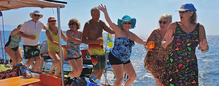 Guests dance and wave on the trimaran's shaded deck with the sea behind them
