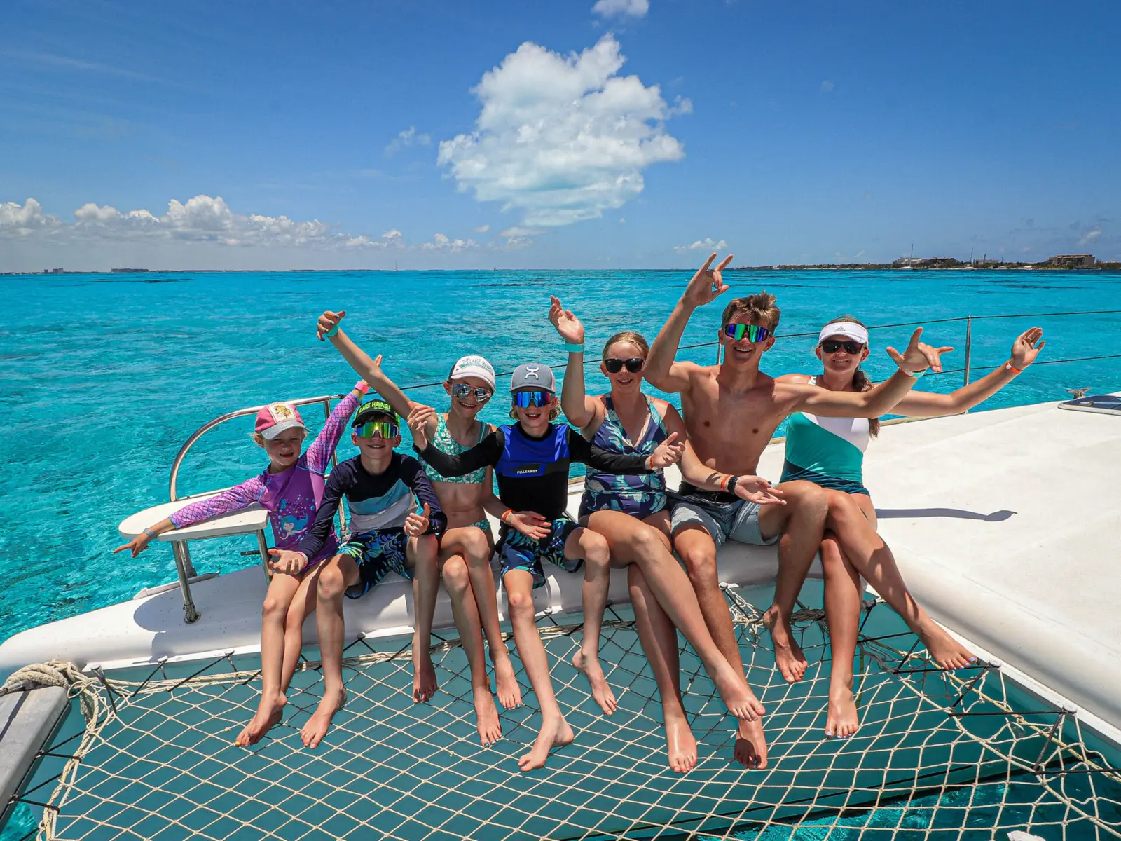 Adults and kids in rash guards waving from the catamaran's net over turquoise water off Cancún