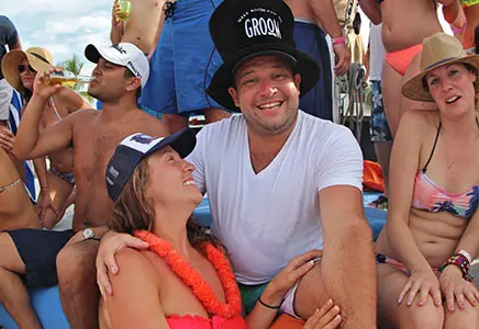 Man in a black top hat reading Groom, celebrating with friends in swimwear, one in a flower lei