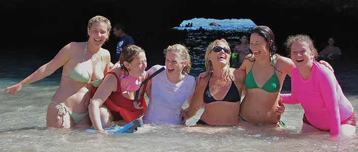 Six women laughing together in the shallows of a hidden beach under a rocky overhang near Puerto Vallarta