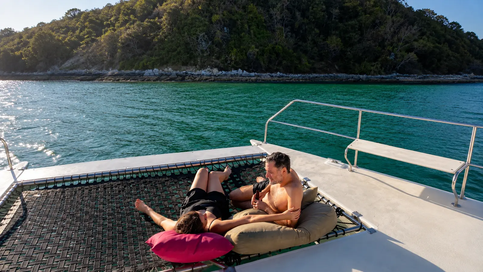 A couple lounges on cushions on the trimaran's bow netting, a forested island across the water