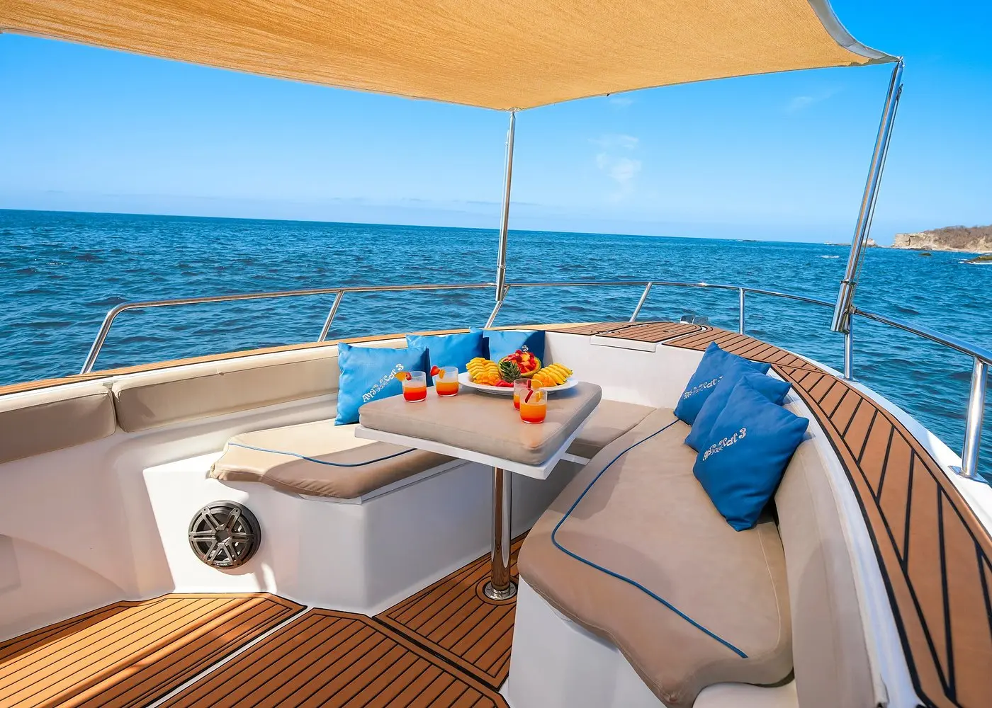 Shaded bow lounge of a center-console boat with teak decking, blue cushions and a table of fruit and drinks