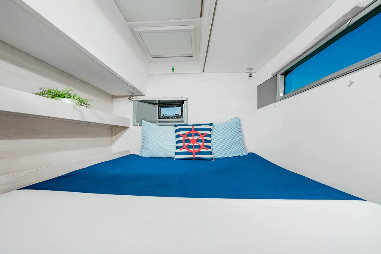 Catamaran cabin with a double bed in navy linens, nautical pillows and a hull window