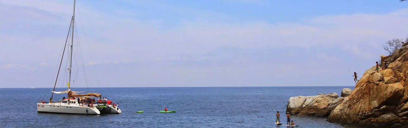 Sailing catamaran anchored off a rocky point near Puerto Vallarta while guests kayak and explore