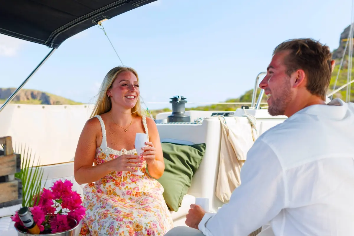 Couple laughing over drinks in the shaded cockpit of a sailing yacht beside a basket of pink flowers