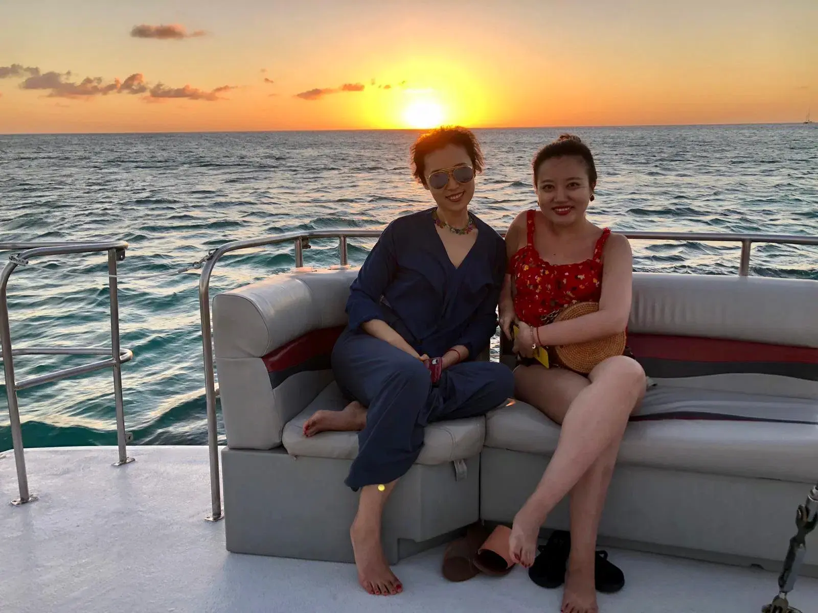 Two women sit together on a cushioned bench aboard the catamaran with the sun low over the ocean