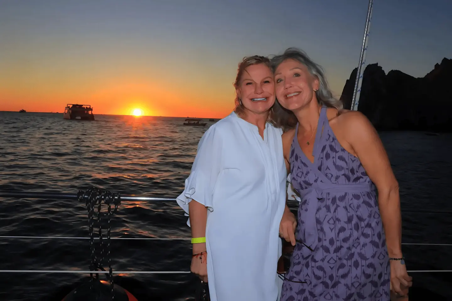 Two women smile on the catamaran at dusk, the sun dipping into the sea beside a dark rocky outcrop