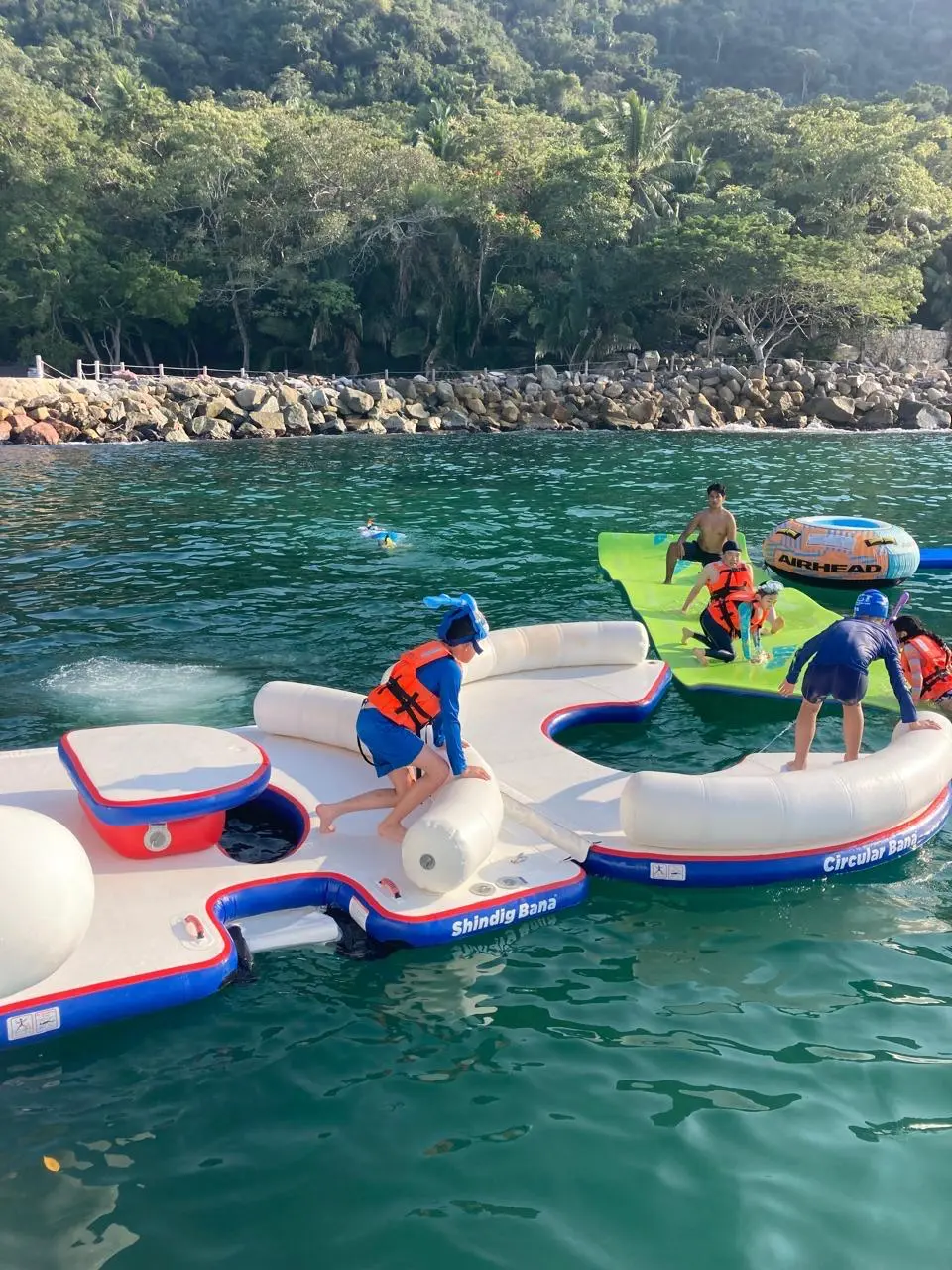 Guests in life jackets climb onto a red and white inflatable platform near a jungle-covered Banderas Bay shore