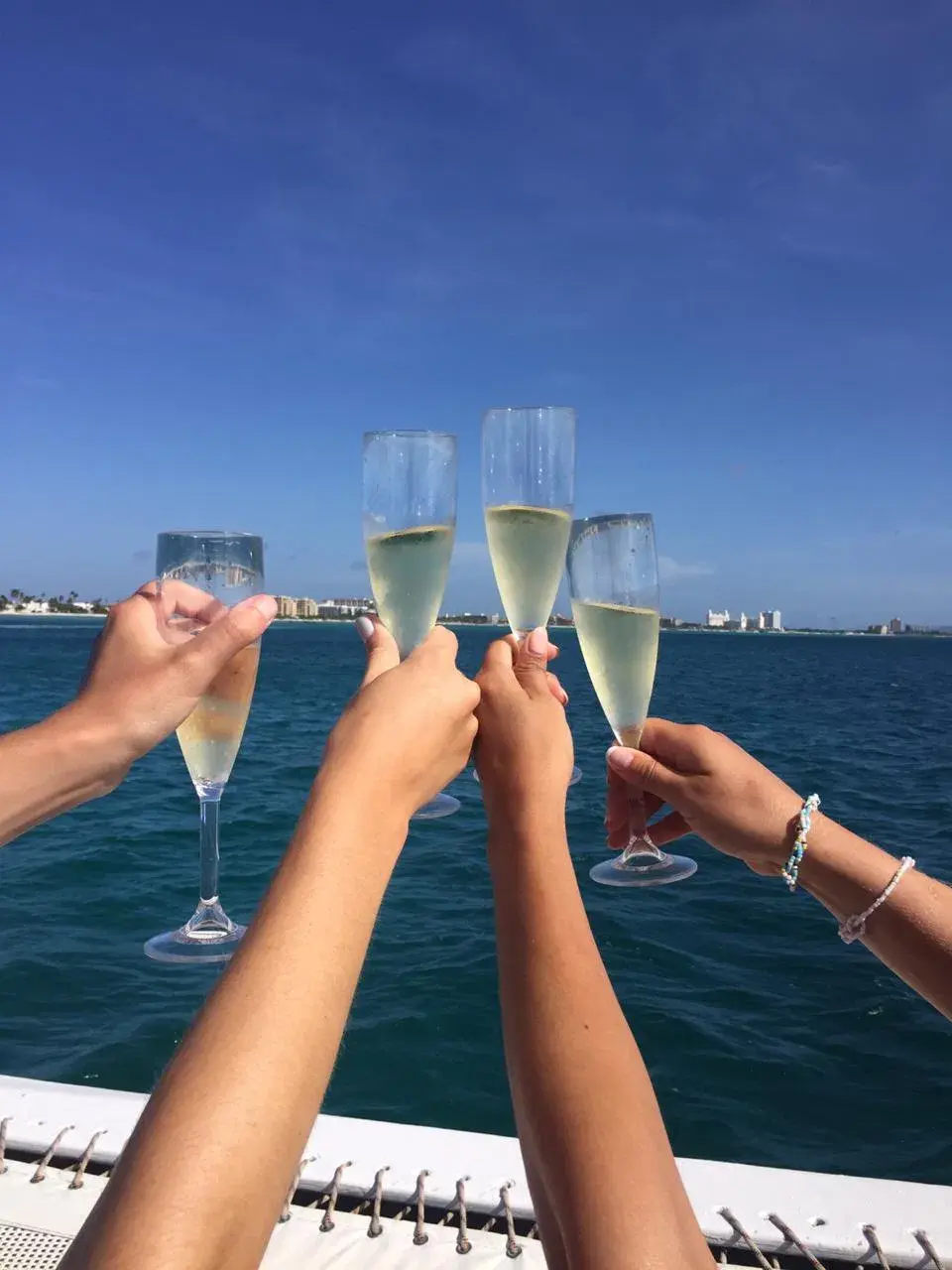 Four hands clink glasses of bubbly in a toast over blue water on a sunny day